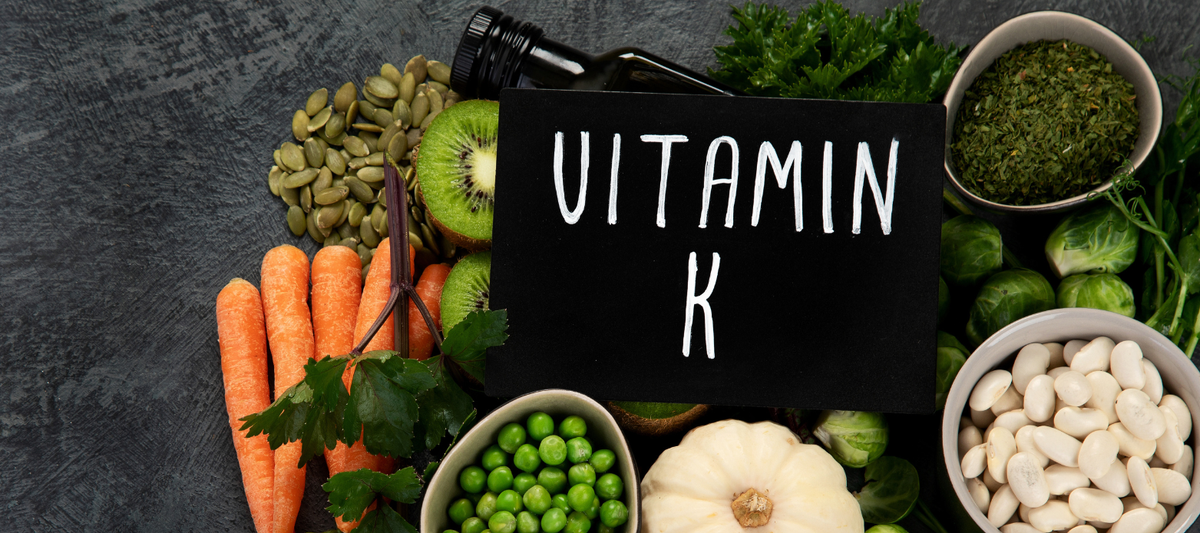 Tafel mit der Aufschrift 'Vitamin K' umgeben von Kürbiskernen, Kiwi, Karotten, Erbsen, weißen Bohnen und grünem Gemüse