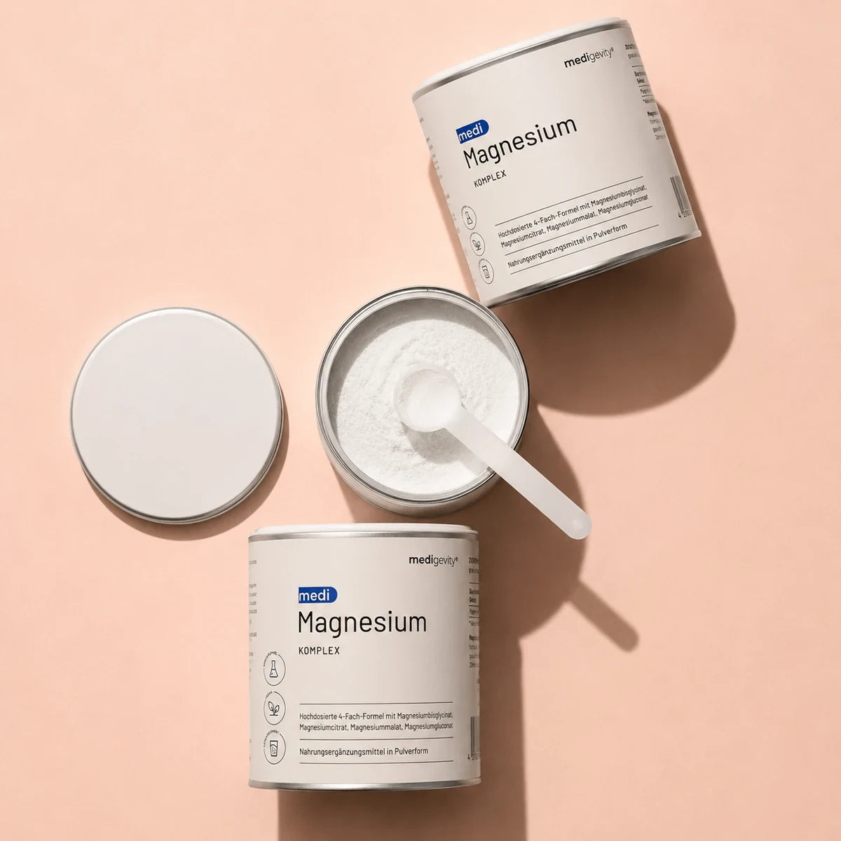 medi Magnesium Komplex Pulverdosen, eine geöffnet mit Dosierlöffel, auf rosafarbenem Hintergrund
