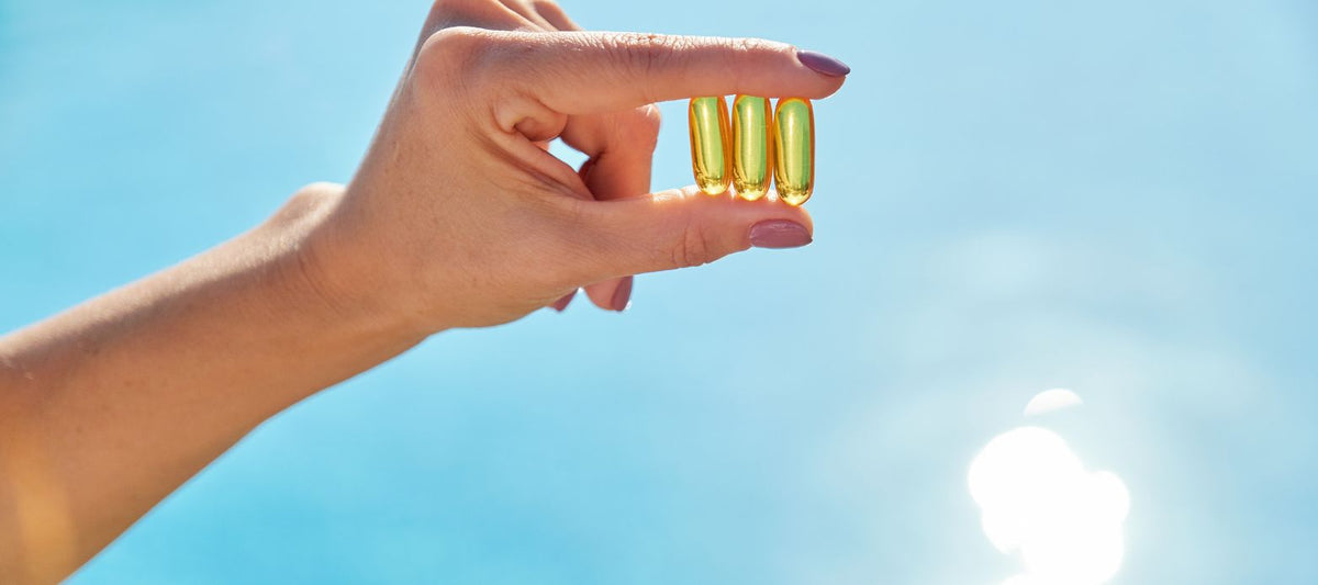 Hand hält drei Omega-3-Kapseln vor blauem Himmel mit Sonnenlicht