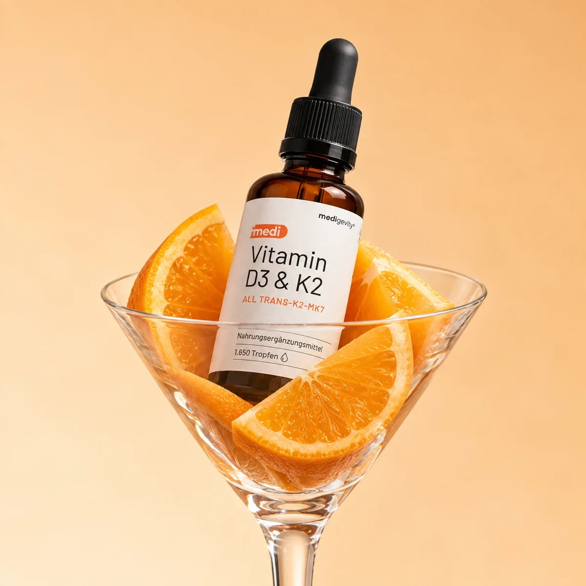 medi Vitamin D3 & K2 Tropfenflasche (1.650 Tropfen) in einem Cocktailglas mit Orangenspalten vor warmem Orangehintergrund