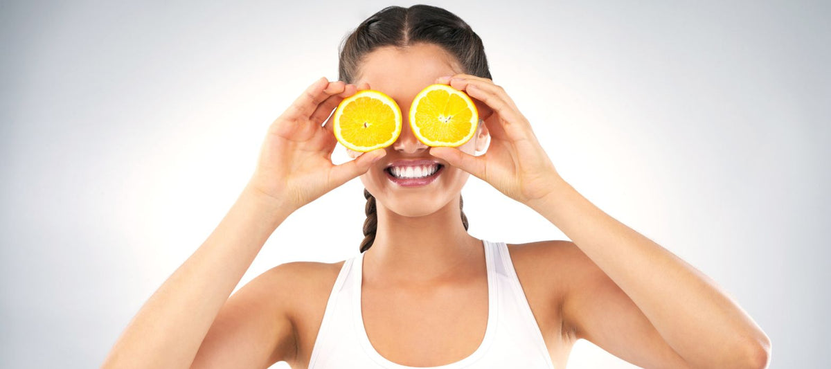 Lächelnde Frau hält sich zwei Orangenscheiben vor die Augen – heiteres Symbolbild für Vitamin C aus Zitrusfrüchten